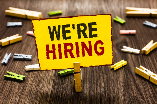 istock Handwriting text writing We re are Hiring. Concept meaning Advertising Employment Workforce Placement New Job Clothespin holding yellow paper note several clothespins wooden floor. 1069879358