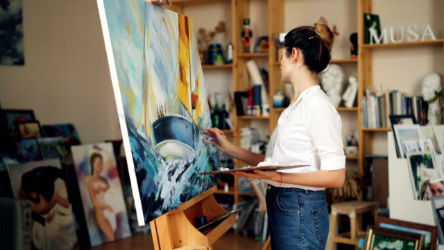 Good-looking young woman in casual clothing is painting in workroom then looking at picture, evaluating her work and smiling enjoying beautiful image.