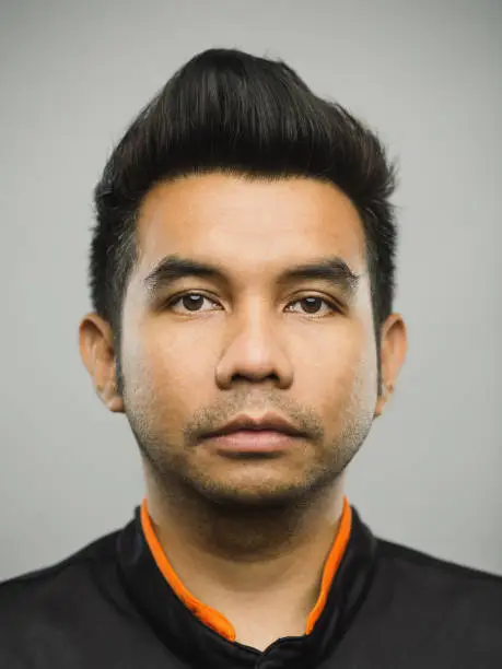 Close up portrait of asian young man with blank expression against gray background. Vertical shot of real malaysian man staring in studio with black hair and quiff. Photography from a DSLR camera. Sharp focus on eyes.