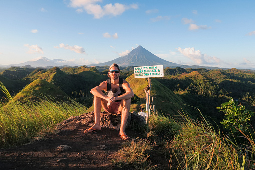 A wide-view shot of a male tourist sitting down near the chocolate hills in the Philippines, the chocolate hills can be seen as well as a mountain in the distance, followed by a clear blue sky.