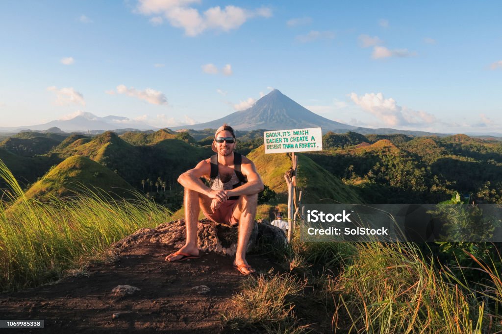 Tourist Visiting Chocolate Hills A wide-view shot of a male tourist sitting down near the chocolate hills in the Philippines, the chocolate hills can be seen as well as a mountain in the distance, followed by a clear blue sky. Hiking Stock Photo