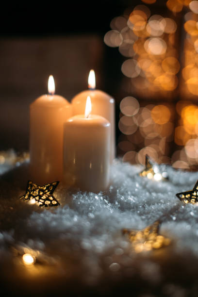 A close up studio shot of a centrepiece decoration for Christmas and New Year holiday celebration. The centrepiece features several white fine candles burning standing on white cupcake liner on a grey ceramic plate. The plate stands on a white paper dolly. The plate also contains Christmas ornaments in silver colour. A white fringe ribbon decorates the arrangement. Defocused white Christmas lights at the background. Main colours are white, silver colored and grey.
