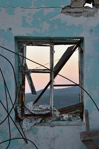 Broken window in abandoned house with tangled rusty wires and distant mountain view during the blue hour.