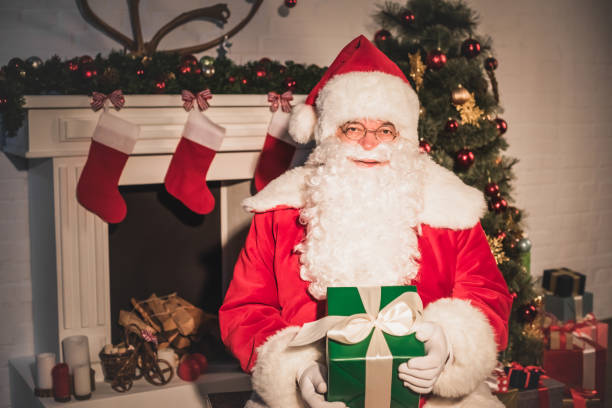 istock santa claus holding christmas gift and looking at camera while sitting near fireplace 1070053010