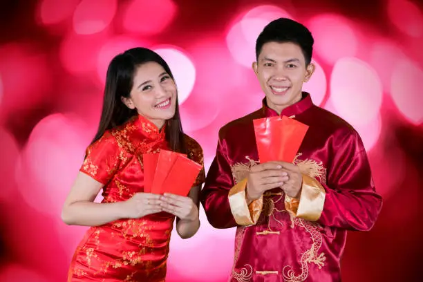 Picture of happy couple wearing cheongsam clothes while holding envelopes and standing with blurred sparkling light background