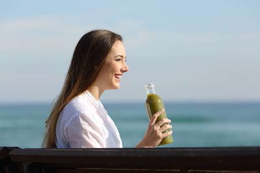 Side view portrait of a woman holding a vegetable juice sitting on a bench on the beach