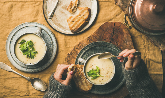 Autumn, Winter home dinner. Flat-lay of Fall warming celery cream soup and female hands in sweater with spoon over linen tablecloth, top view. Comfort food, vegetarian, healthy and slow food concept