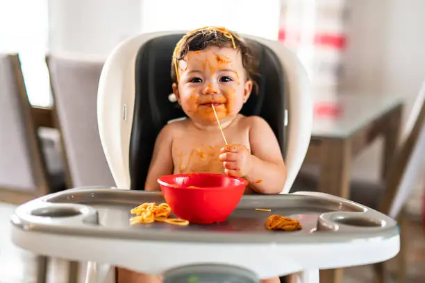 Photo of child girl, eating spaghetti for lunch and making a mess at home in kitchen