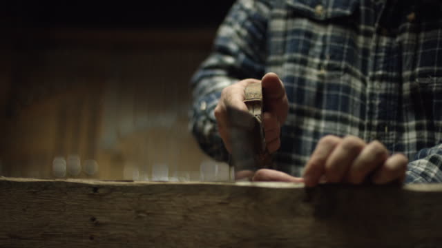 A Fifty-Something Male Woodworker in a Plaid Shirt Uses an Antique Hand Saw to Saw a Red Oak Board in a Workshop