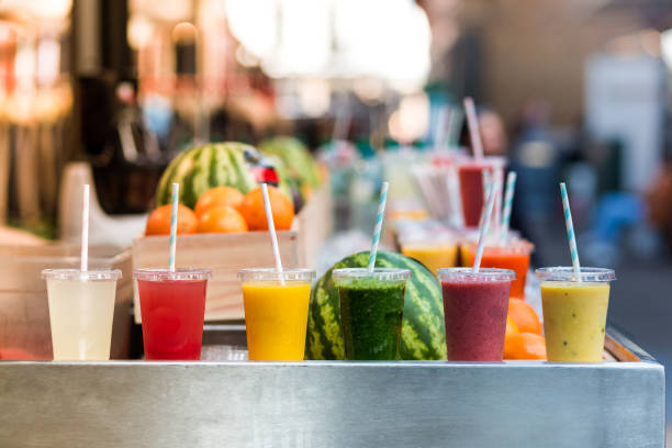 Close up of fresh fruit smoothies and juices in a row for sale on vegetarian market stall Close up color image depicting freshly made fruit juices and smoothies on display in a row and for sale at a food and drink market in London, UK. Selective focus on the plastic cups containing the fresh smoothies. In the background people are blurred out of focus. Room for copy space. fruit juice stock pictures, royalty-free photos & images