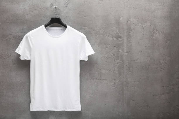 Front side of male white cotton t-shirt on a hanger and a concrete wall in the background Front side of male white cotton t-shirt on a hanger and a concrete wall in the background. T-shirt without print and copyspace for your text on right side coathanger stock pictures, royalty-free photos & images