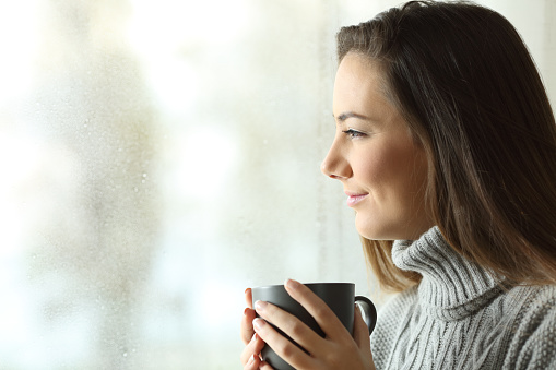 Happy woman looking at rain holding coffee through the window of home or hotel
