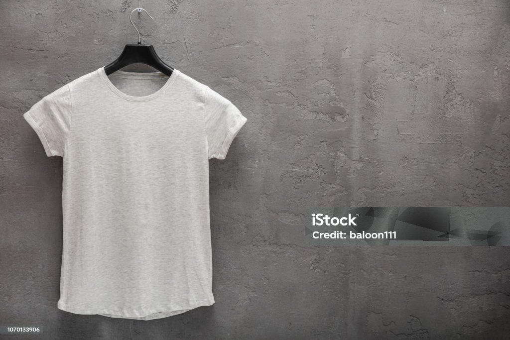 Front side of female grey melange cotton t-shirt on a hanger and a concrete wall in the background Front side of female grey melange cotton t-shirt on a hanger and a concrete wall in the background. T-shirt without print and copyspace for your text on right side T-Shirt Stock Photo