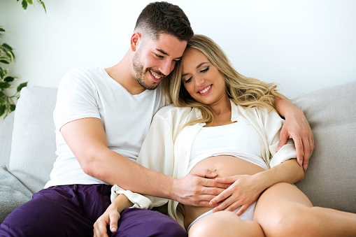 Shot of handsome young man touching belly of beautiful pregnant woman while lying on the sofa at home.