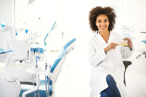 Young african american female dentist sitting in a dental office, holding denture and toothbrush and showing how to brush teeth correctly. She's smiling and looking at camera.