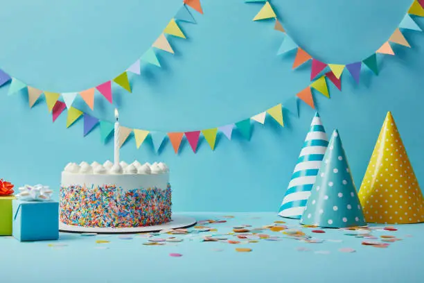 Photo of Delicious birthday cake, gifts, party hats and confetti on blue background with bunting