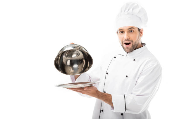 istock excited young chef taking of serving dome from plate and looking at camera isolated on white 1070153612