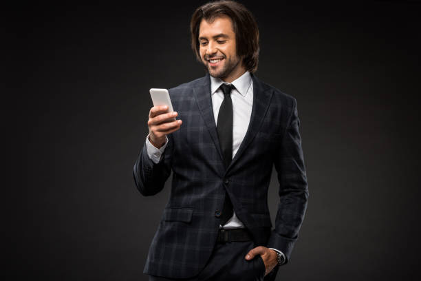 istock smiling young businessman standing with hand in pocket and using smartphone isolated on black 1070175942