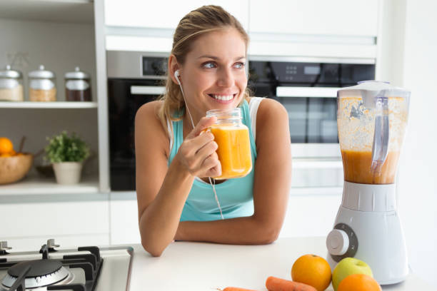 beautiful sporty young woman looking sideways and drinking vegetable detox juice in the kitchen at home. - orange smoothie imagens e fotografias de stock