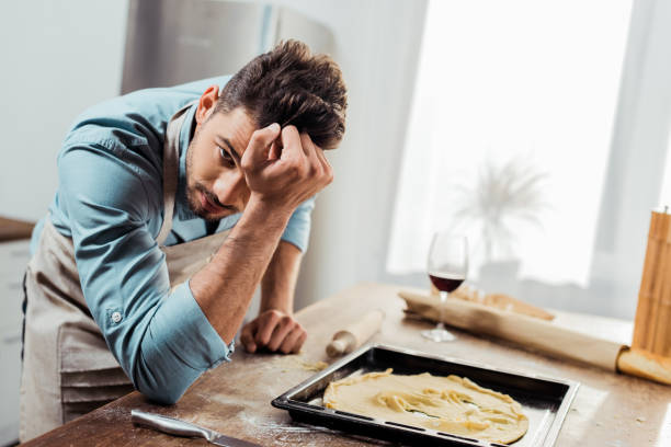 istock upset young man in apron looking at camera while leaning at table with spoiled dough on baking tray 1070231526