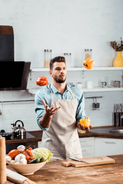 istock handsome young man in apron juggling with fresh peppers in kitchen 1070232100