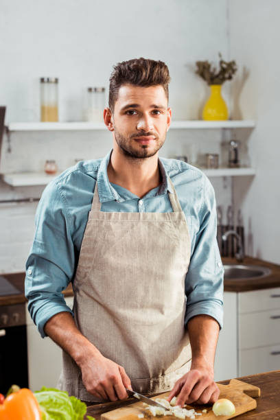 istock handsome young man in apron chopping onion and looking at camera in kitchen 1070232498