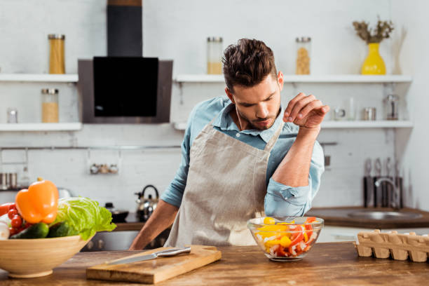 istock handsome young man in apron adding salt to vegetable salad while cooking in kitchen 1070232658