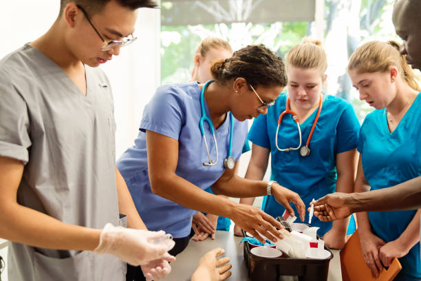Multi-ethnic group of students in college nursing class. Multi-ethnic group of College students in nursing class. Woman professor is explaining how to do blood tests. Everybody is wearing a uniform. They are in a mock hospital room with mannequin arm on table. Horizontal indoors waist up shot. medical student photos stock pictures, royalty-free photos & images
