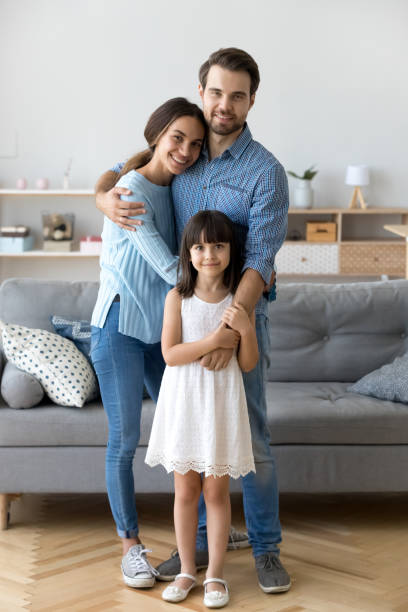 Family standing in living room embracing looking at camera Full length diverse multi-ethnic family married couple wife husband little daughter embracing standing together in living room smiling looking at camera at new modern home feels happy and satisfied adoption photos stock pictures, royalty-free photos & images