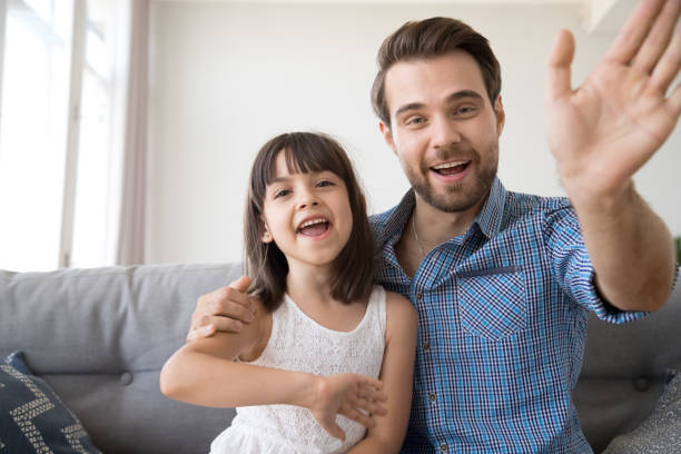 Happy father and daughter wave hands looking at camera Head shot portrait of cheerful smiling diverse family looking at camera. Young male sitting on couch at home with daughter have video call using computer, wave hands greeting or say goodbye to friend divergent movie review stock pictures, royalty-free photos & images