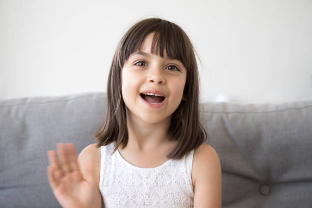 Head shot portrait little girl wave hand looking at camera Small girl sitting on couch alone at home. Head shot portrait little adorable daughter waving hand looking at camera smile saying hello or goodbye. First acquaintance and online communication concept divergent movie review stock pictures, royalty-free photos & images