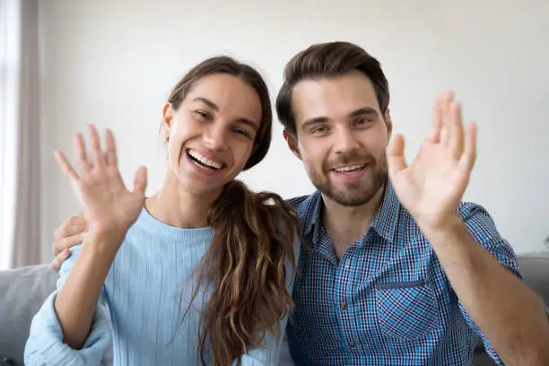 Head shot married couple sitting on sofa at home. Portrait wife and husband waving hands looking at camera smiling saying hello greeting or goodbye. First acquaintance or online communication concept
