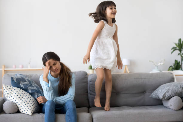 istock Tired mother has headache little daughter jumping on couch 1070262164