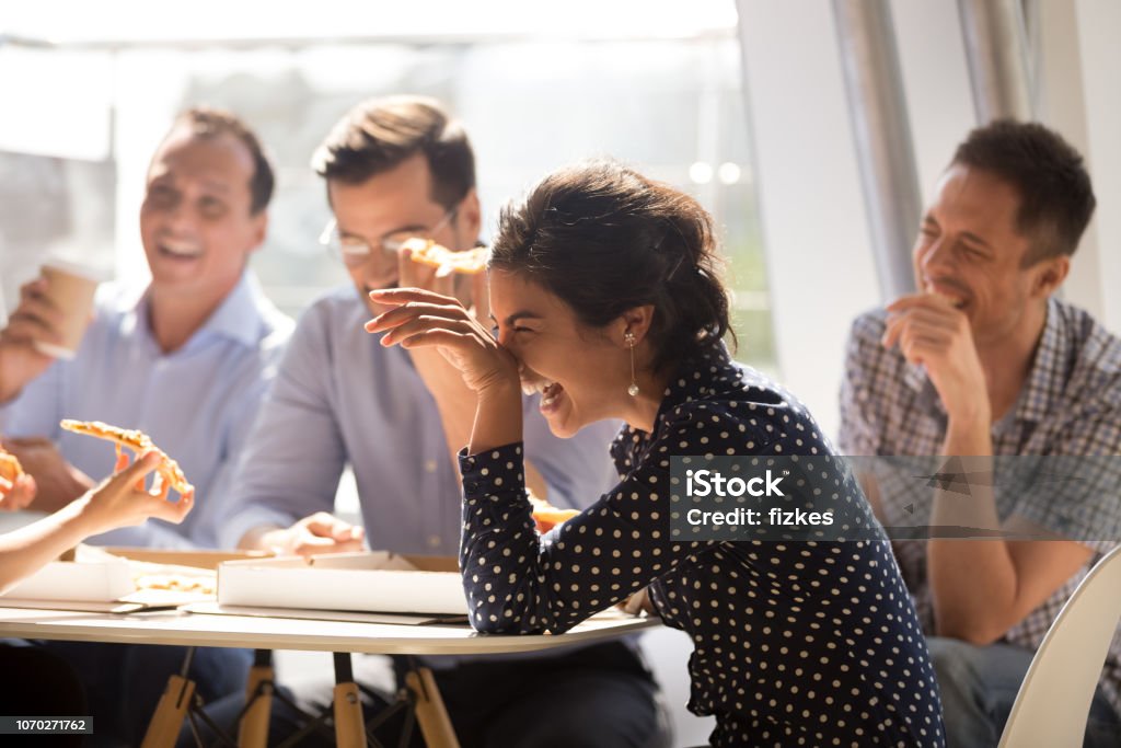 Indian woman laughing eating pizza with diverse coworkers in office Indian woman laughing at funny joke eating pizza with diverse coworkers in office, friendly work team enjoying positive emotions and lunch together, happy colleagues staff group having fun at break Laughing Stock Photo