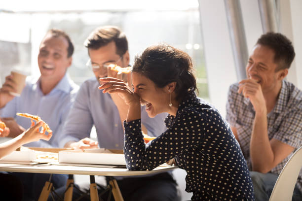 Indian woman laughing eating pizza with diverse coworkers in office Indian woman laughing at funny joke eating pizza with diverse coworkers in office, friendly work team enjoying positive emotions and lunch together, happy colleagues staff group having fun at break humor stock pictures, royalty-free photos & images