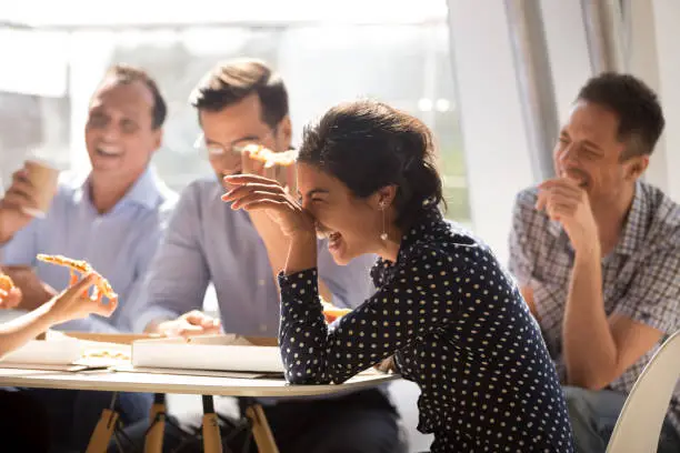 Photo of Indian woman laughing eating pizza with diverse coworkers in office