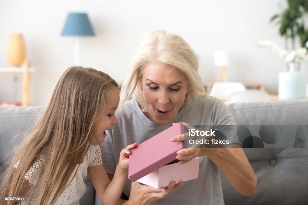 Excited grandmother and granddaughter unpacking birthday gift together Excited granddaughter open birthday present together with granny, spend time together at home having fun, happy grandmother and grandchild celebrate making surprise, unpacking gift box Gift Stock Photo