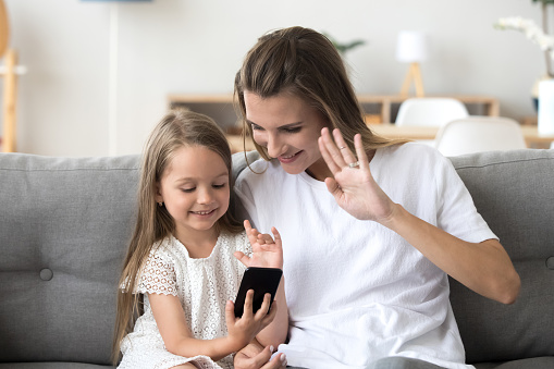 Happy mom and daughter make video call on smartphone talking with dad or relatives, smiling little girl and mother waving at cellphone, having skype conversation from home. Kids and technology concept