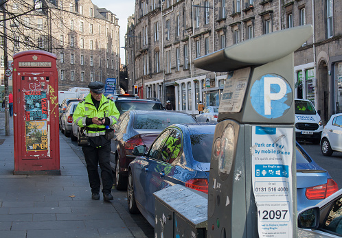 Edinburgh, Scotland. 11/8/18. Parking attendant wearing uniform and checking cars in a street of Edinburgh, Scotland, with parking meter in the foreground and british red phone box in background