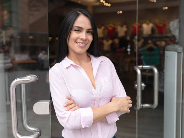 amérique latine femelle (la) propriétaire d’un magasin de vêtements se penchant sur la porte d’entrée souriant à la caméra - boutique store owner latin american and hispanic ethnicity photos et images de collection