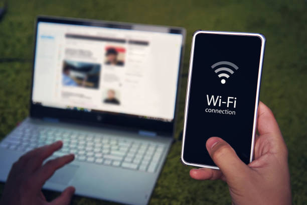 istock Smartphone with wifi connection generic icon on the screen hold by man on the background of laptop display and keyboard. Man uses Wi-Fi hot spot to get to domestic internet access. Wireless gadgets 1070356876