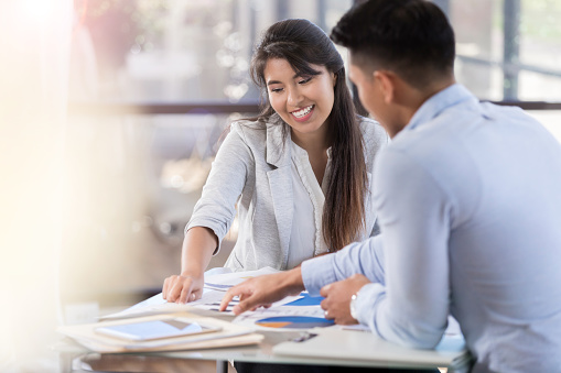 A young man and woman sit at a table covered in paperwork. They point at particular papers and smile.