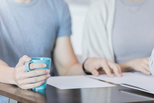 Two people sit at a table and read through papers. They hold onto their mug and follow the lines with their fingers. The camera is zoomed in.