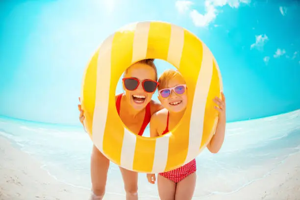 smiling modern mother and child in red swimsuit on the seashore looking through yellow inflatable lifebuoy