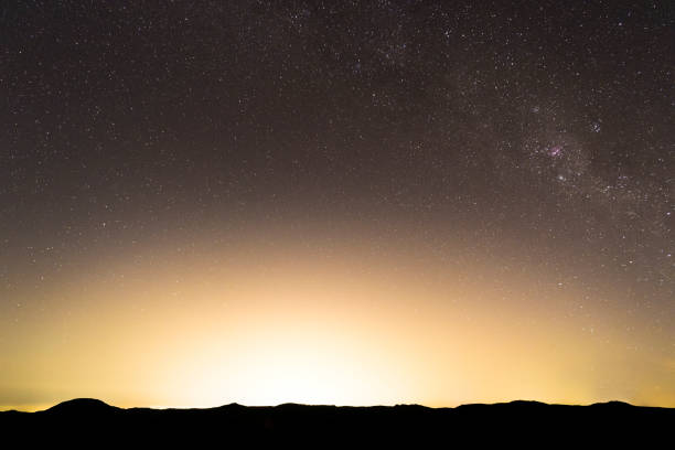the moon and stars stand above the lights of the city of santa fe with the sandia mountains far on the horizon.  a cellphone tower sticks up into the sky and contrasts the scenery.  horizontal composition taken at christmas time in santa fe, new mexico.  