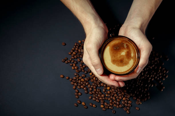 cup of hot coffee in the hands with scattered roasted grains on the table - fotografia de stock