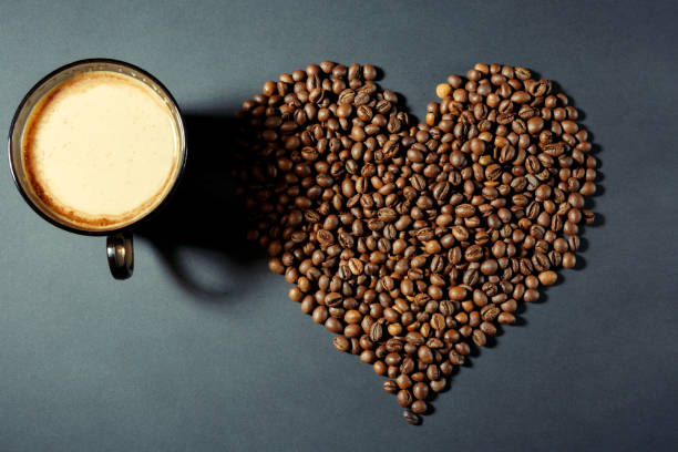 roasted grains in the shape of a heart and a cup of aromatic coffee on a table in a dark vein - fotografia de stock