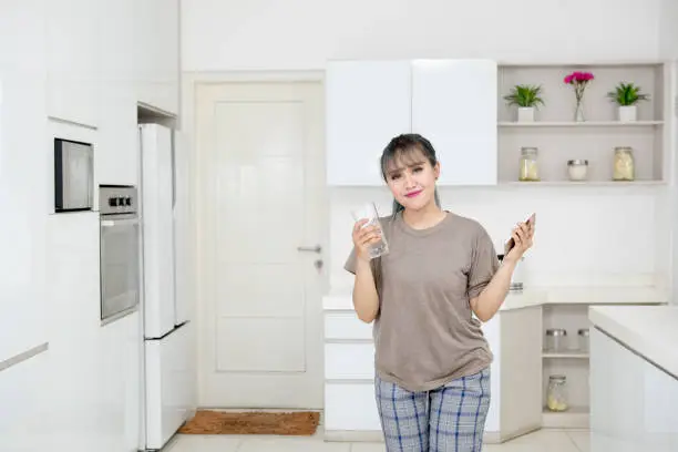 Pretty girl holding a mobile phone and glass of water while standing in the kitchen