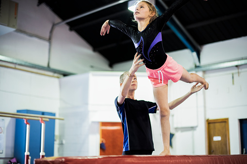 A 9 year old girl learning gymnastics. Her coach, a mature woman in her 40s, is helping her on the balance beam.