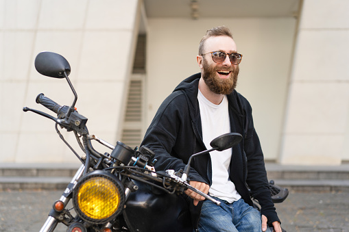 Joyful biker resting on motorbike. Bearded young man in sunglasses and black casual jacket sitting on motorcycle and laughing. Leisure concept